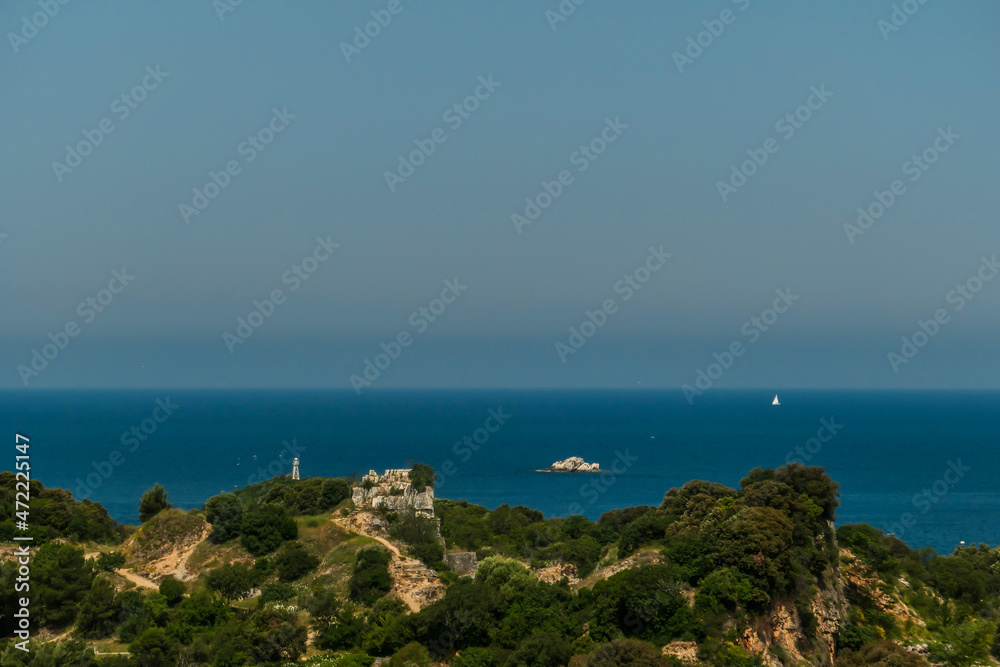 A view on the ruins of a castle seen from a hill above the ruins. The castle was located at the headland, directly by the sea. There are few boats crossing the sea. Clear and sunny day.