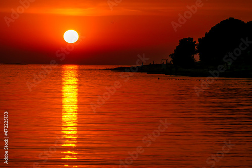 Romantic sunset by a beach. The sun sets over the horizon. The sun beams reflecting in the calm sea waters. There is an island on the side. Few birds flying around. The sky turns yellow and orange