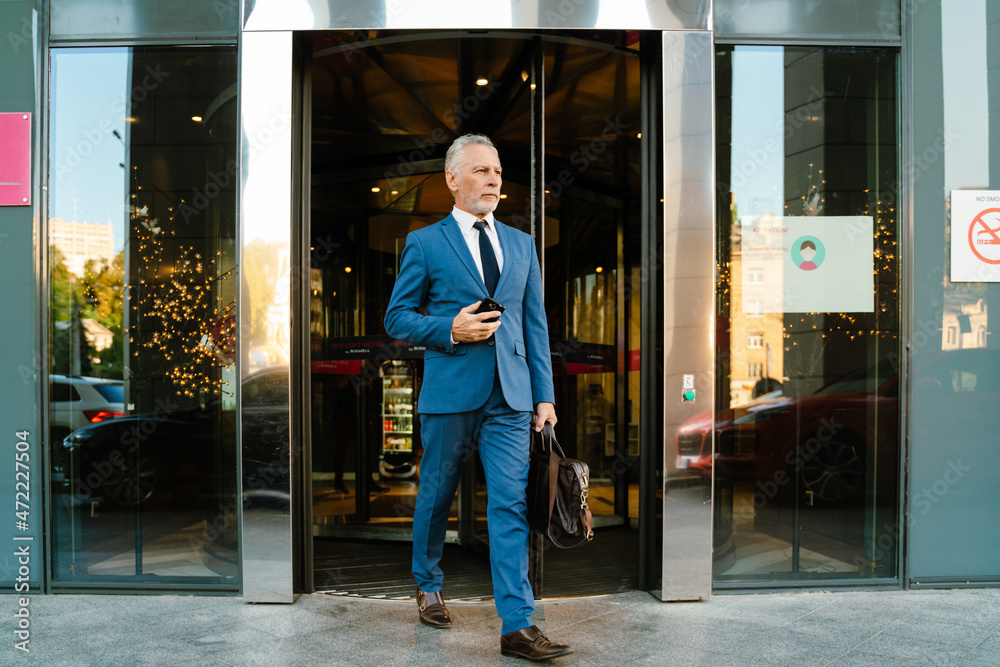 Senior man dressed in suit using mobile phone while coming out door Senior man dressed in suit using mobile phone while coming out door