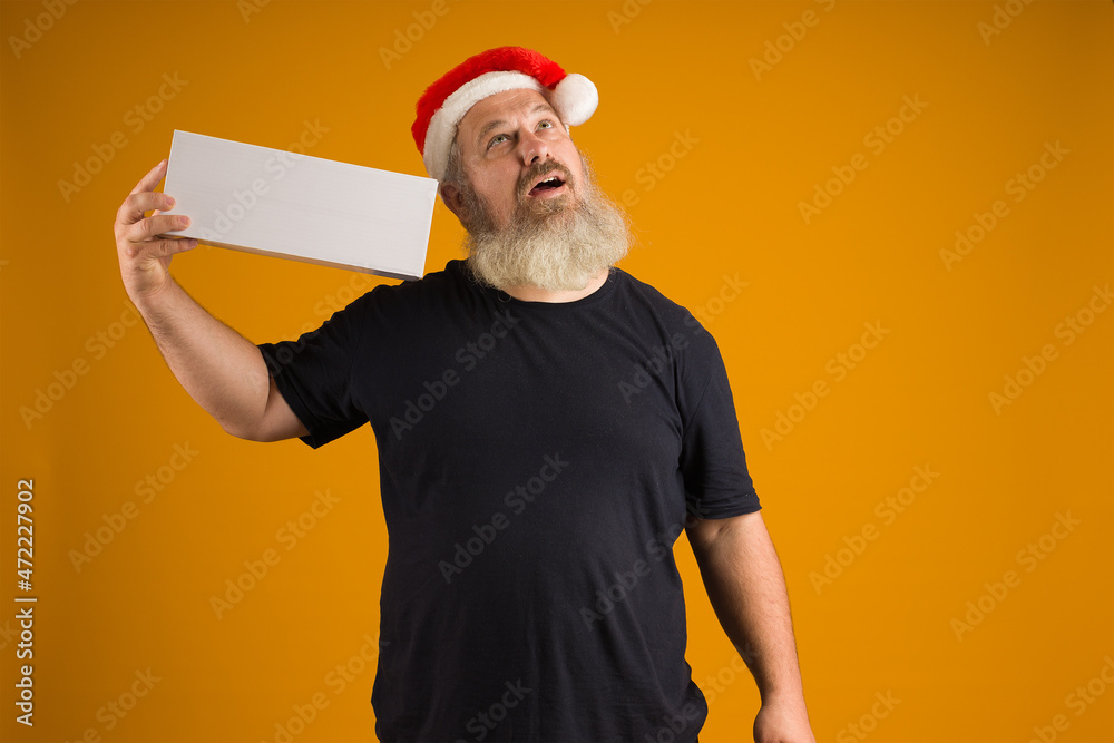 man in santa claus hat with a beard, black t-shirt, holding a white box on his shoulder and looking up against a yellow background, mockup