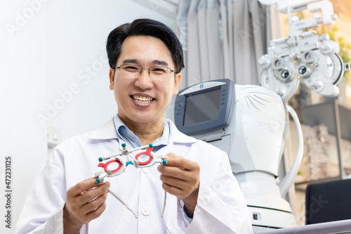 Asian man optometrist smiling and holding adjustable optical trial lenses frame in clinic. Eye test and optometry in optical shop concept.