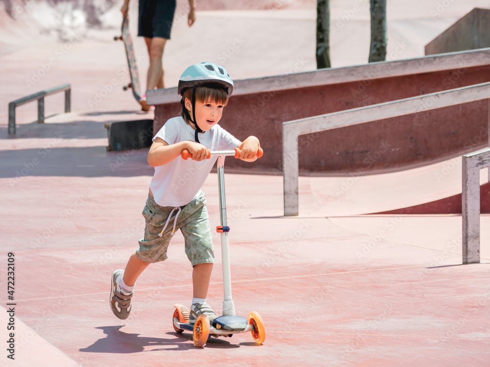 Little boy rides kick scooter in skate park. Special concrete bowl structures in urban park. Training to skate at summer.