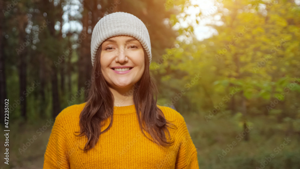 Attractive woman tourist wearing warm knitted hat and orange sweater smiles to camera spending time in suburban green forest close view Attractive woman tourist wearing warm knitted hat and orange sweater smiles to camera spending time in suburban green forest close view