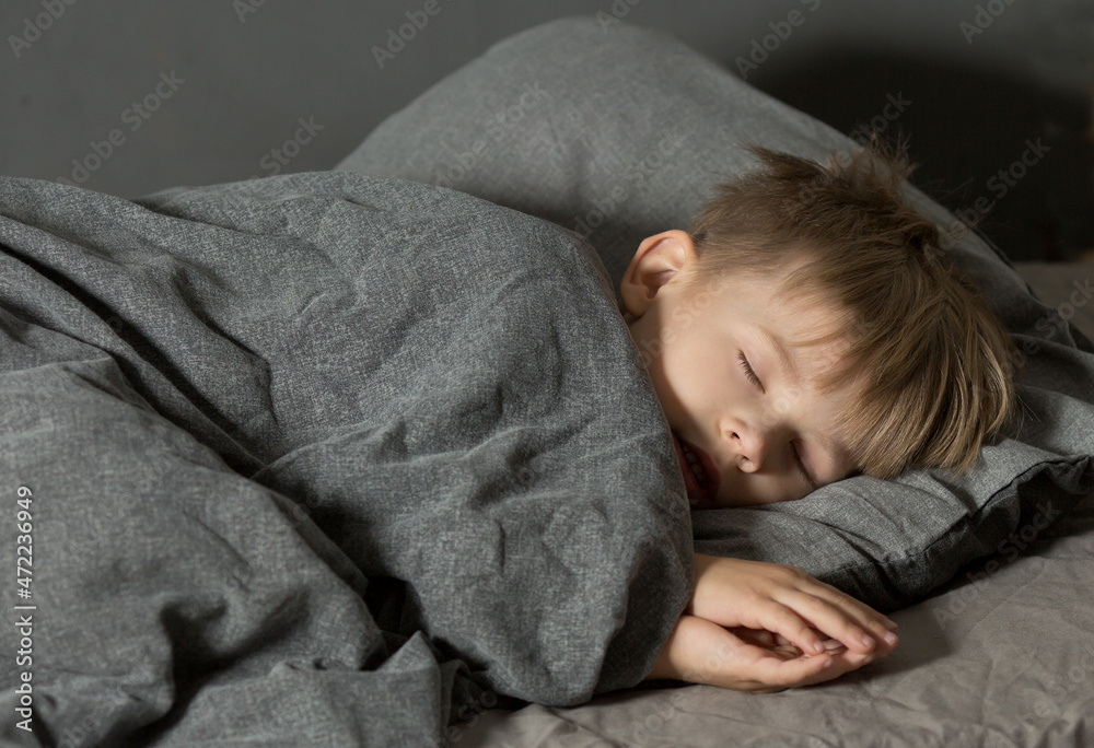 a 5-year-old preschooler boy sleeps in bed on a pillow under a blanket. Grey dark background a 5-year-old preschooler boy sleeps in bed on a pillow under a blanket. Grey dark background