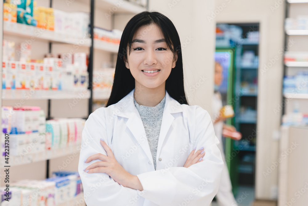 Closeup photo of asian female confident successful young pharmacist druggist in white medical coat standing with arms crossed at the cash point desk in pharmacy drugstore