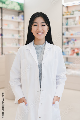 Vertical photo of asian female confident young pharmacist druggist in white medical coat looking at camera at the cash point desk in pharmacy drugstore
