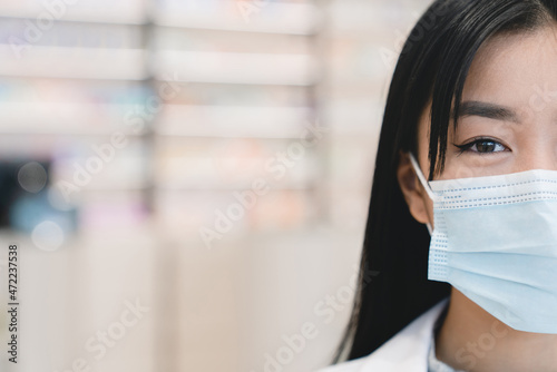 Closeup cropped shot of asian female young pharmacist druggist in white medical coat and protective face mask against Covid19 looking at camera in pharmacy drugstore