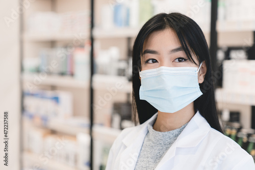 Pandemic, lockdown. Asian female young pharmacist druggist in white medical coat and protective face mask against Covid19 looking at camera in pharmacy drugstore