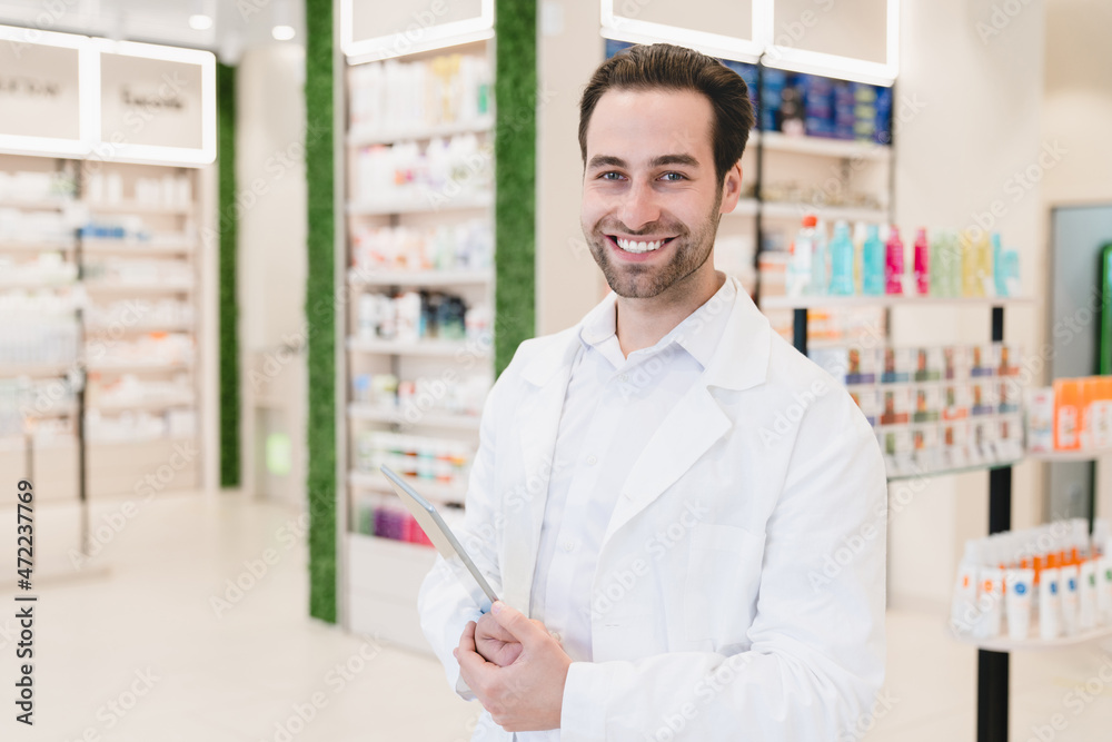 Vertical portrait of caucasian male young pharmacist druggist in white medical coat holding clipboard with side effects, active substance, prescriptions standing at pharmacy drugstore