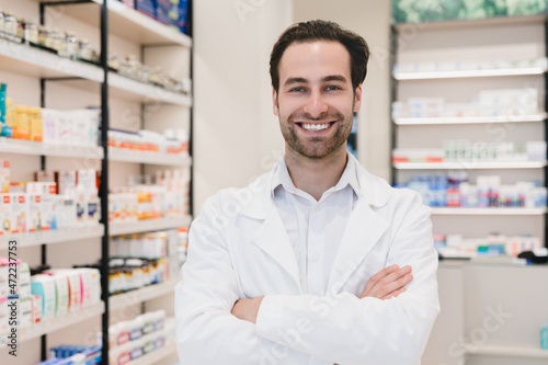 Closeup photo of male caucasian confident successful young pharmacist druggist in white medical coat standing with arms crossed at the cash point desk in pharmacy drugstore