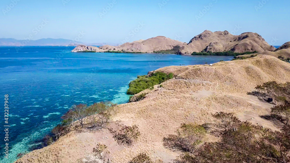 A drone shot of a paradise island in Komodo National Park, Flores, Indonesia. Brownish island turns into white sand beach and further into turquoise and navy blue sea. Steep slopes of the island