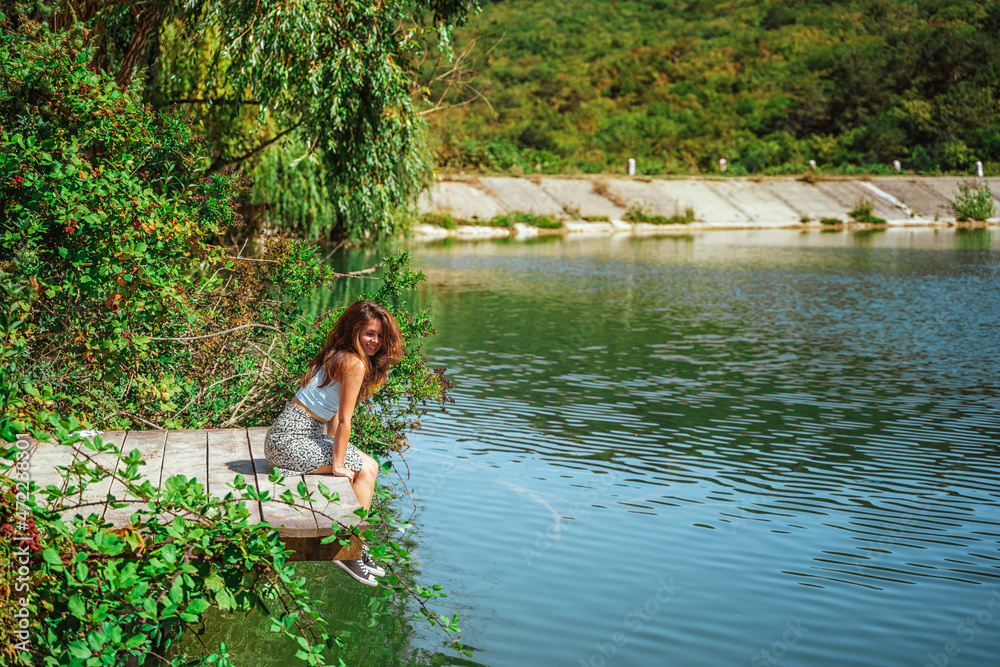 Fototapeta premium A charming young woman in a skirt is sitting on a wooden bridge against the background of nature with a lake in the forest. Travel, freedom, the concept of an active lifestyle.