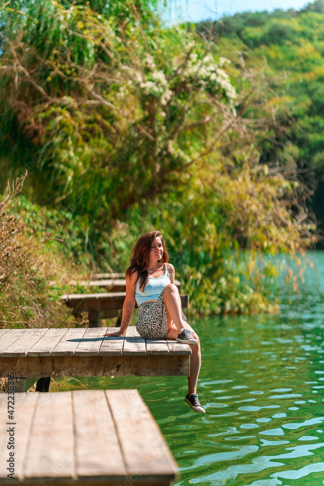 A charming young woman in a skirt is sitting on a wooden bridge against the background of nature with a lake in the forest. Travel, freedom, the concept of an active lifestyle. A charming young woman in a skirt is sitting on a wooden bridge against the background of nature with a lake in the forest. Travel, freedom, the concept of an active lifestyle.