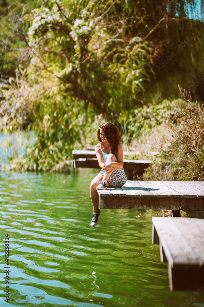 Fototapeta premium A charming young woman in a skirt is sitting on a wooden bridge against the background of nature with a lake in the forest. Travel, freedom, the concept of an active lifestyle.