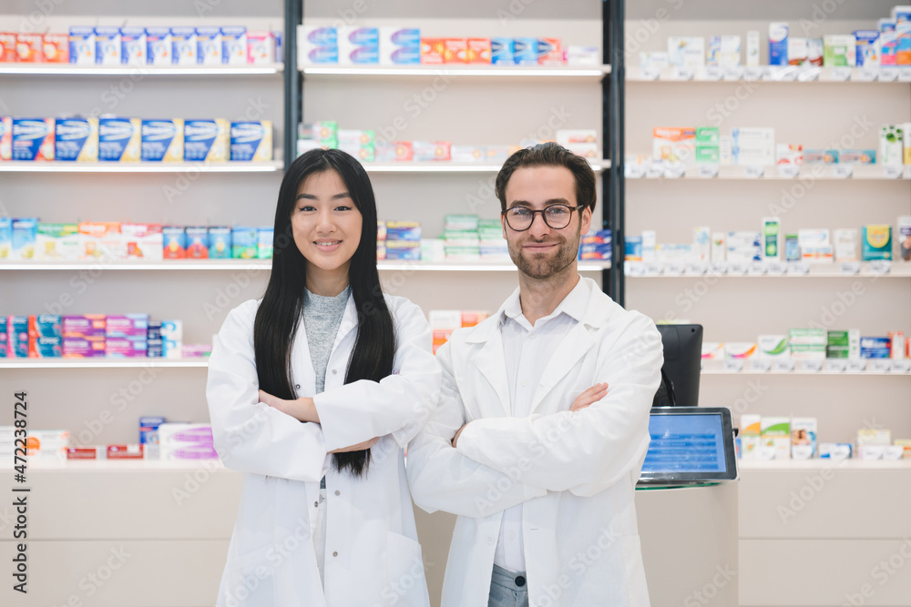 Two confident successful young pharmacists druggists in white medical coats standing with arms crossed at the cash point desk in pharmacy drugstore