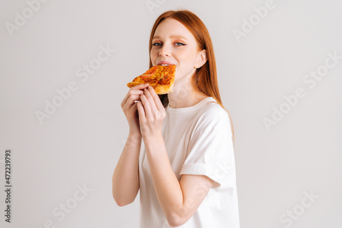 Studio portrait of cheerful happy young woman with appetite to eat delicious pizza looking at camera standing on white isolated background. Pretty redhead female eating tasty meal.