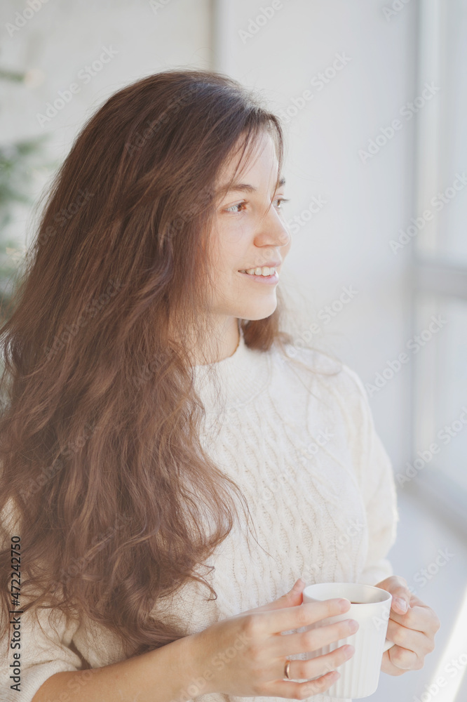 Beautiful young woman drinking coffee or tea near christmas tree in the living room at home closeup Beautiful young woman drinking coffee or tea near christmas tree in the living room at home closeup