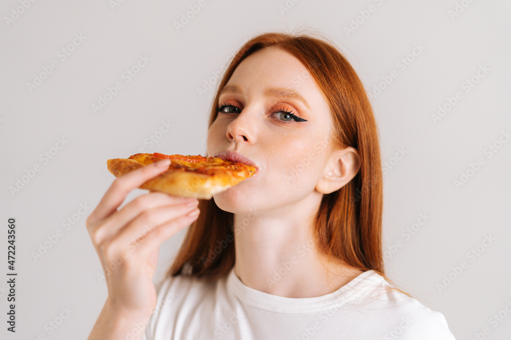 Close-up face of happy attractive young woman with appetite eating delicious pizza looking at camera standing on white isolated background. Pretty redhead female eating tasty meal.