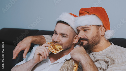 Handsome gay male couple with santa hats eating pizza and watching movie on christmas eve. High quality photo
