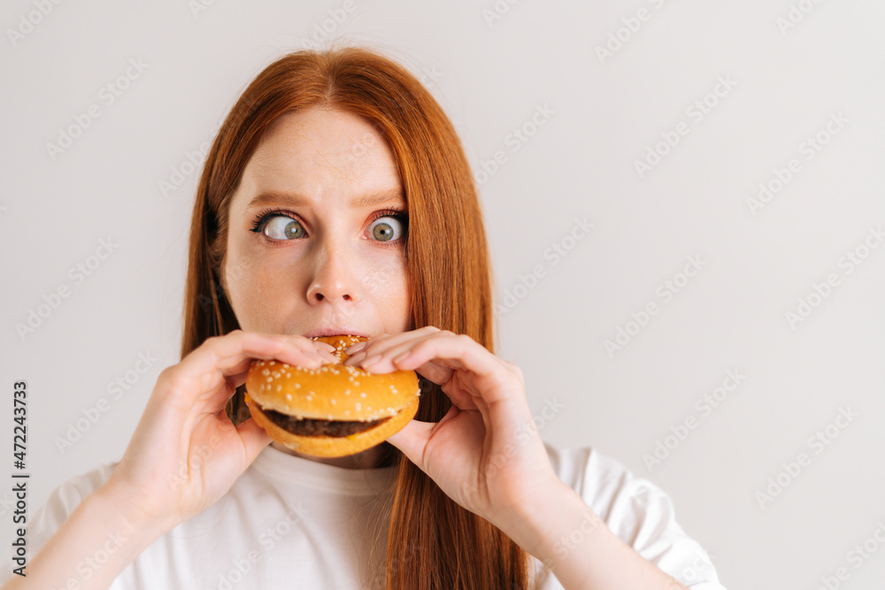 Close-up of funny fooling around young woman with strabismus pleasing bite of appetizing delicious burger looking at camera, standing on white isolated background in studio, front view, portrait. Close-up of funny fooling around young woman with strabismus pleasing bite of appetizing delicious burger looking at camera, standing on white isolated background in studio, front view, portrait.