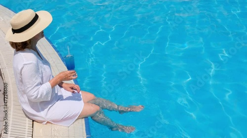 luxury senior woman dangling her legs in an outdoor swimming pool holding a blue refreshing cocktail wearing a straw hat. Summer rest. All inclusive