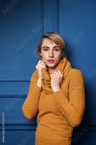 Studio portrait of a young woman in yellow pullover on blue wall background.