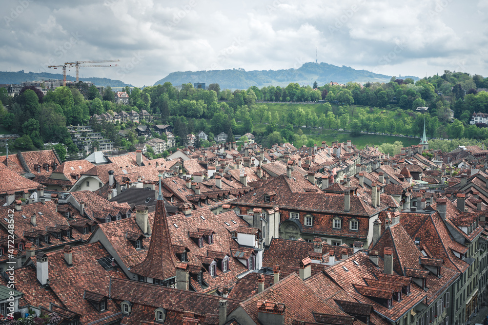 A view of the cityscape of Bern, Switzerland with stone rooftops, streets below, and the farmer's market - a cloudy spring day with green trees and hills in the background
