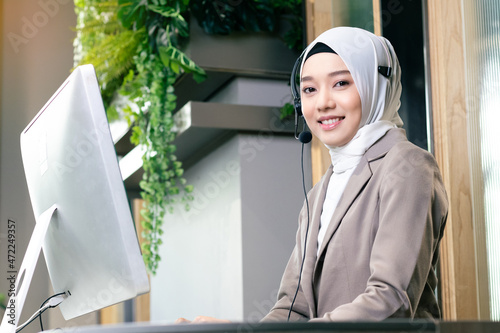 Customer service or call center agent with headset working on support hotline in office, using computer. Young Muslim woman with hijab work as a mature positive agent or receptionist looking at camera