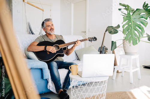 Elderly guitarist performing music on sofa near laptop