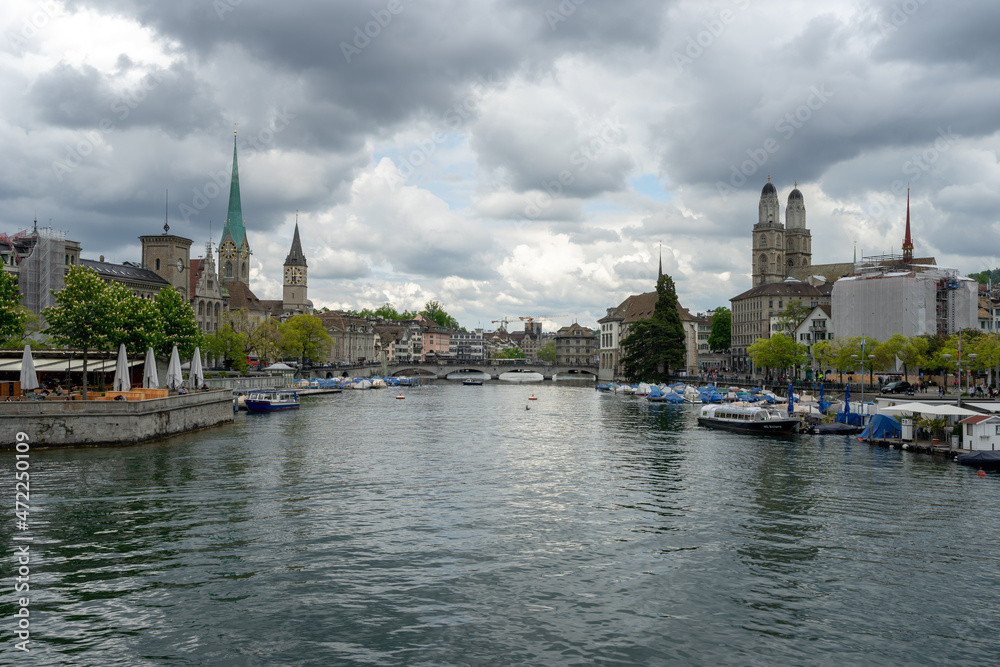 The Limmat River divides the cityscape of Zurich, Switzerland on a cloudy spring afternoon - the spires of churches and buildings rise and boats are docked on the water