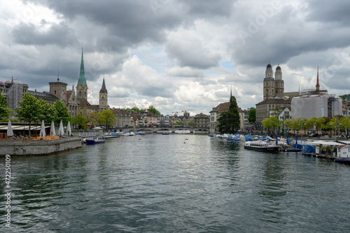 The Limmat River divides the cityscape of Zurich, Switzerland on a cloudy spring afternoon - the spires of churches and buildings rise and boats are docked on the water © Sitting Bear Media