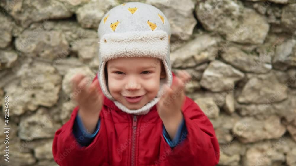 close-up Portrait of European little preschool boy in gray knitted hat red jacket smiling look at camera. Emotionally child laughing smiling posing cover eyes with hands, waves hello outside Autumn close-up Portrait of European little preschool boy in gray knitted hat red jacket smiling look at camera. Emotionally child laughing smiling posing cover eyes with hands, waves hello outside Autumn