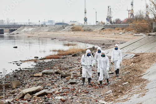 Wide angle view at group of workers wearing hazmat suits walking by water outdoors , toxic waste concept photo
