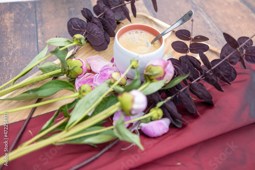 Fototapeta Naklejka Na Ścianę i Meble -  A white Cup of black coffee stands on a wooden background next to pink peonies and other garden flowers and leaves, The concept of village life, Cottagecore, ruralcore, cluttercore. High quality photo