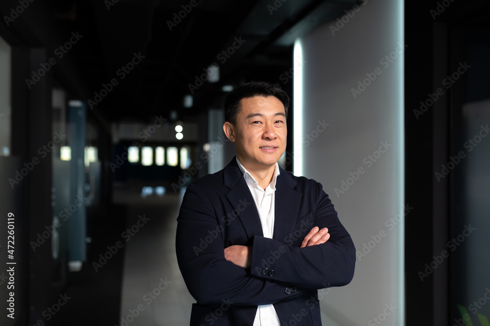 Portrait of a successful Asian manager boss, with crossed arms in a business suit looking pensively at the camera, serious man in the office