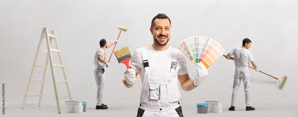 House painter holding a color swatch palette and a brush and other painters painting a wall - obrazy, fototapety, plakaty 