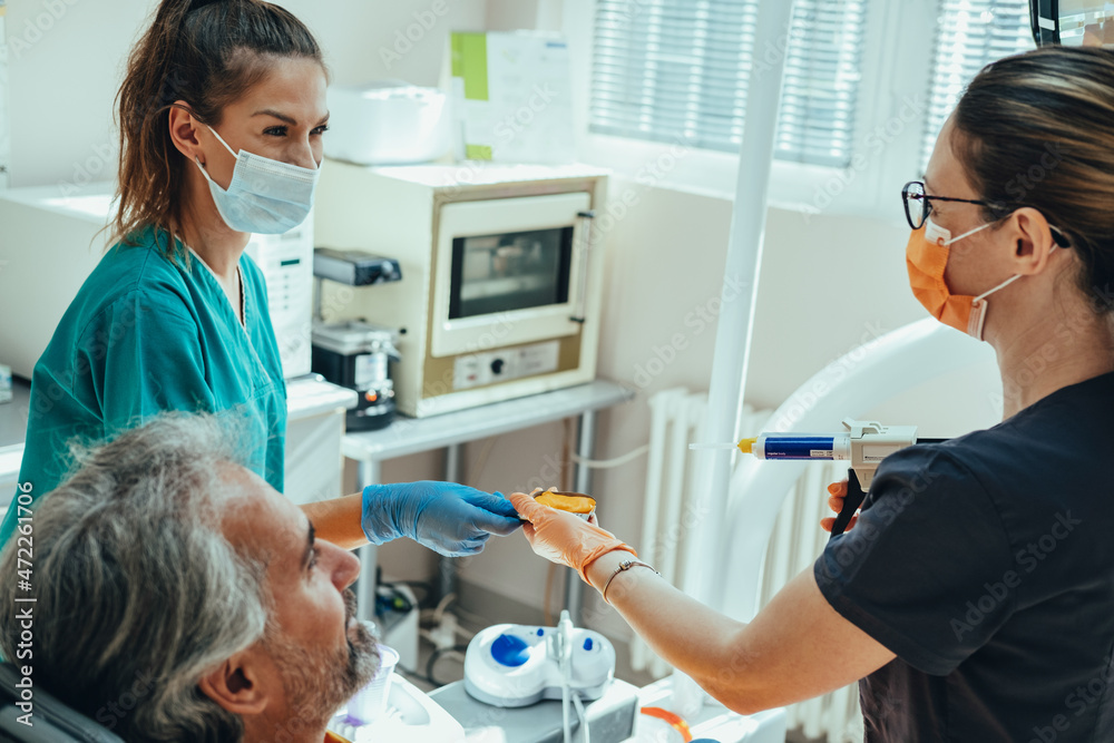 Fototapeta premium Smiling Mature Male Patient Sitting on Dentist's Chair while Female Dentist and Dental Assistant in Protective Face Mask and Gloves Taking Teeth Impression in Dental Clinic