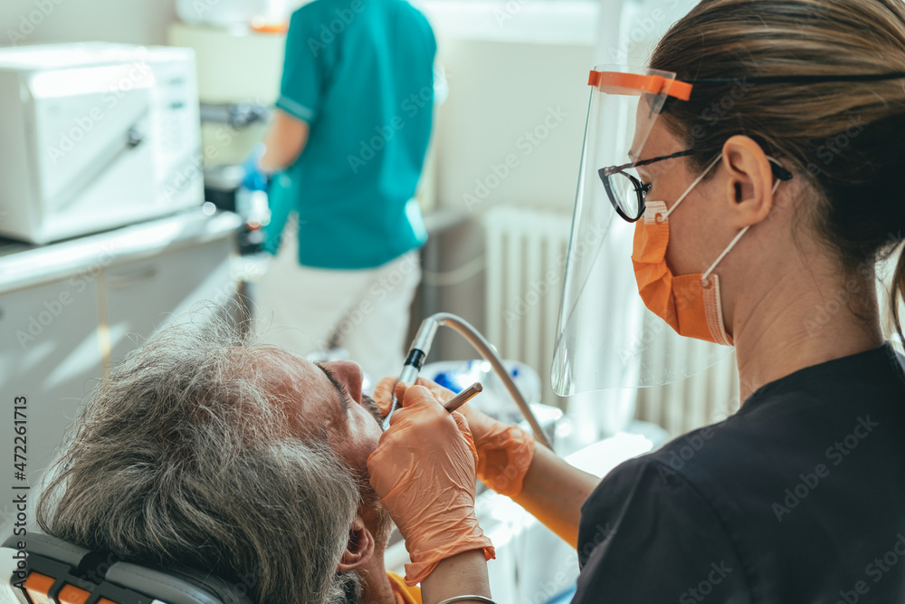 Fototapeta premium Close Up Photo of Woman Dentist in Protective Workwear Using Dental Drill for Treating Dental Cavity on a Male Patient Teeth in a Dental Clinic with Dental Assistant in the Background