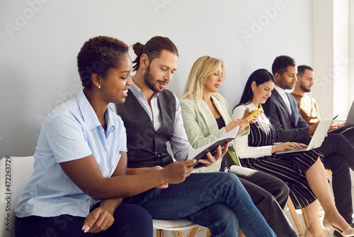Multiethnic people sit in row with gadgets paperwork prepare for interview in company office. Diverse employees or job candidates wait for recruitment talk in queue. Employment and hiring concept.