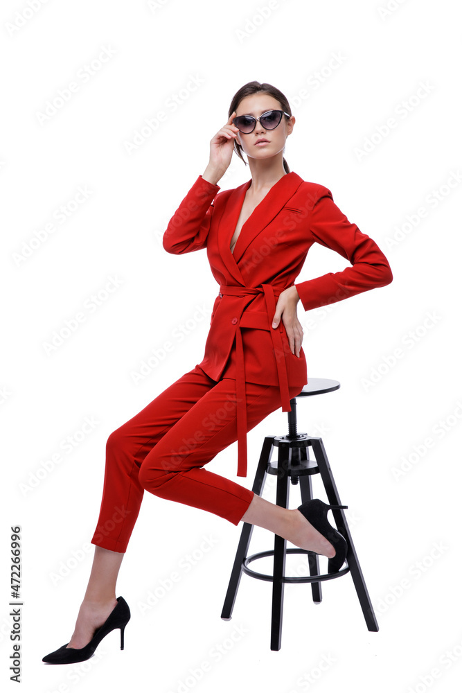 High fashion photo of a beautiful elegant young woman in a pretty red suit, jacket, pants, trousers and sunglasses on white background. Slim figure. Studio Shot. Businesswoman sitting on a black chair