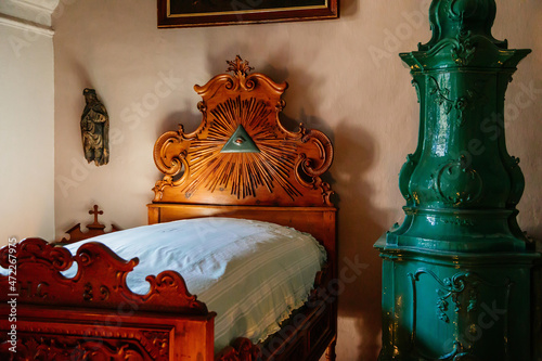 Cervena Lhota, South Bohemia, Czech Republic, 9 October 2021: Castle interior, priest's bedroom, wood carved bed with divine eye and green ceramic tiled stove, mirror in painted frame photo