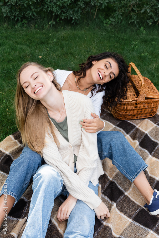 high angle view of joyful lesbian women hugging and laughing on checkered blanket near basket
