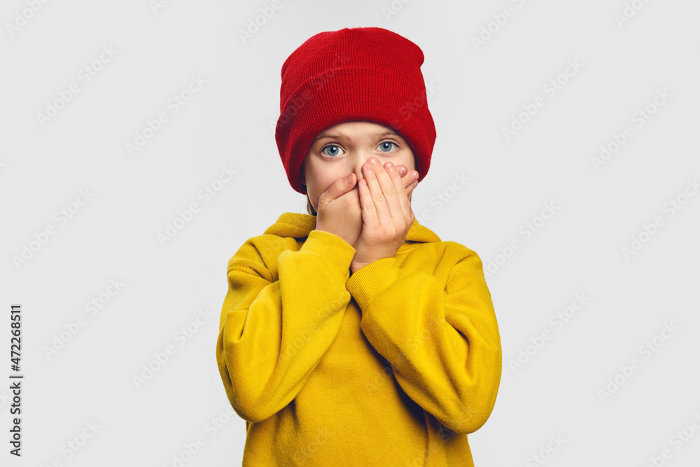 Terrified cute little girl covers mouth with great fear, tries not to cry, has scared shocked expression, wears yellow hoodie and red hat Terrified cute little girl covers mouth with great fear, tries not to cry, has scared shocked expression, wears yellow hoodie and red hat