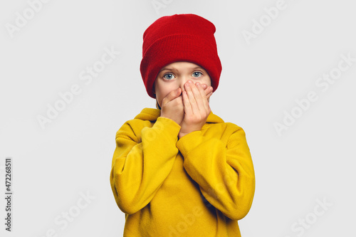 Terrified cute little girl covers mouth with great fear, tries not to cry, has scared shocked expression, wears yellow hoodie and red hat 