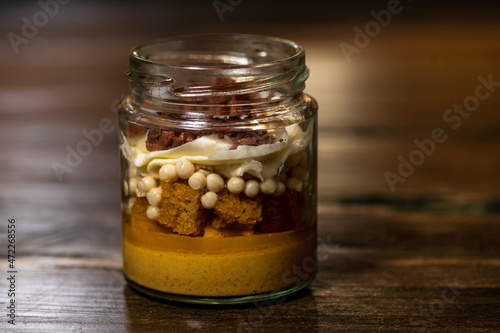 Dessert in a glass tub filled with cream cheese and caramel with white chocolate balls covered with dark chocolate and a crunchy biscuit base