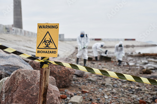 Close up of Warning Biohazard sign at ecological disaster site with people in protective suits, copy space photo