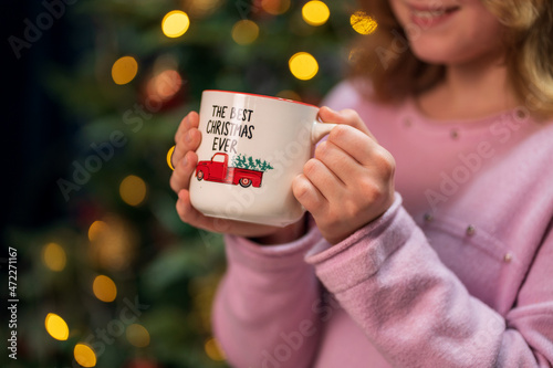 A girl holds a cup with a New Year's image in her hands against a background of Christmas tree lights