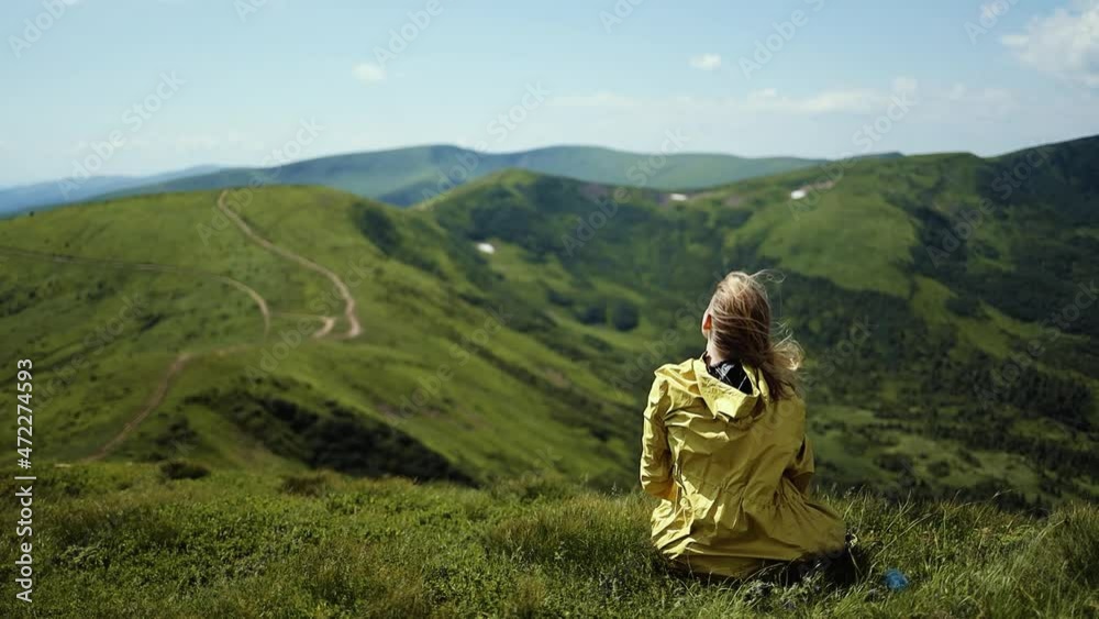 Beautiful blonde woman hiker in a yellow raincoat tourist relaxing on top of mount enjoying view of nature. Back view