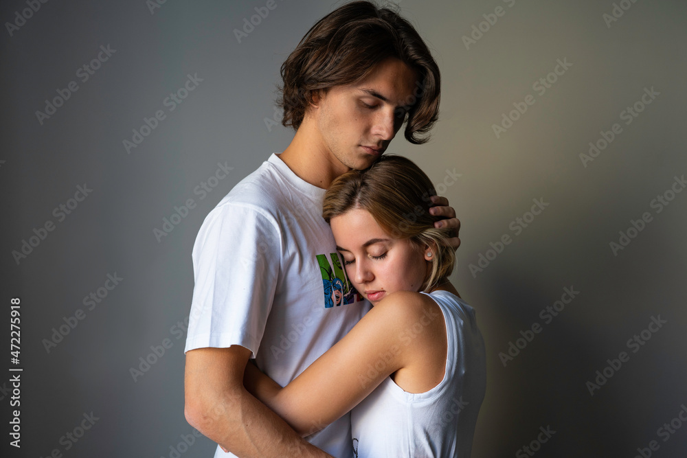 Obraz premium A young couple, a young man and a young blond woman, standing and embracing, he is holding her in hugs with closed eyes both, pressing tenderly her head to his chest, dreaming