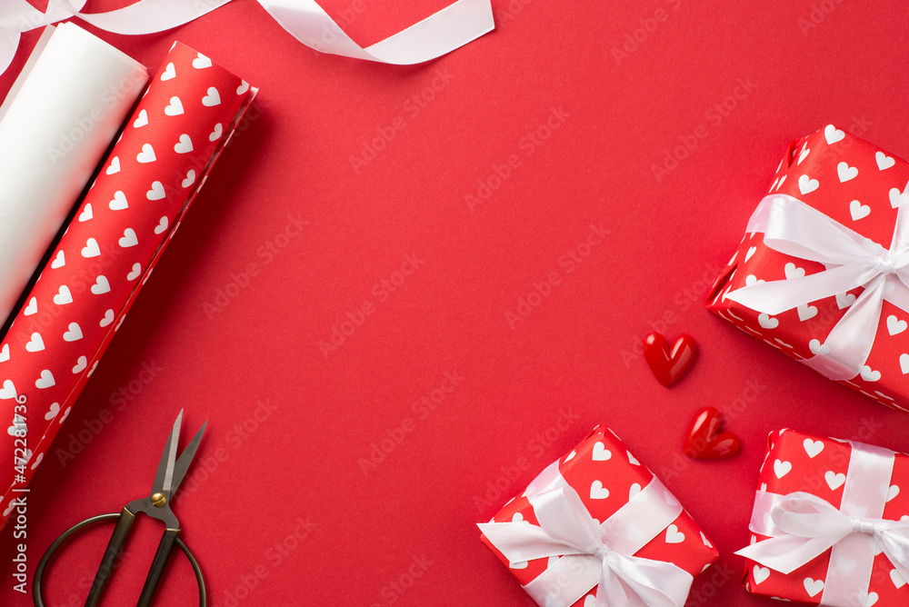 Fototapeta premium Top view photo of gift boxes in red wrapping paper with pattern of hearts and white ribbon bows small hearts rolls of wrapping paper and scissors on isolated red background with copyspace